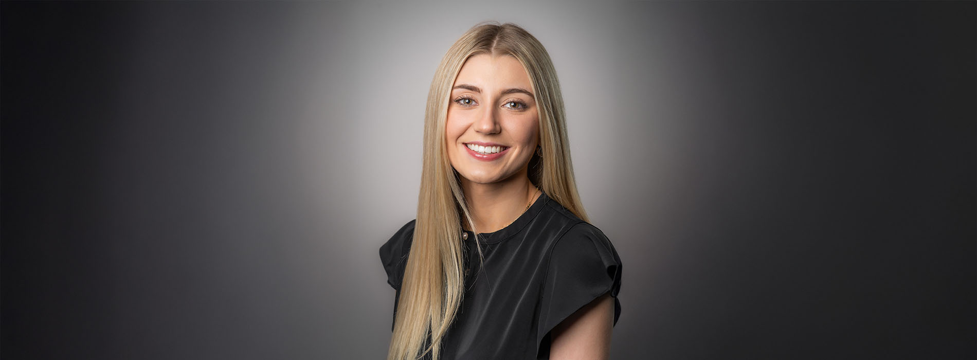 The image shows a woman with blonde hair smiling at the camera against a grey background.